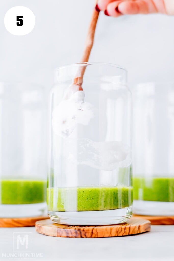 A hand holds a spoon with white cream, about to add it to a glass containing a layer of green liquid; two similar glasses are in the background.