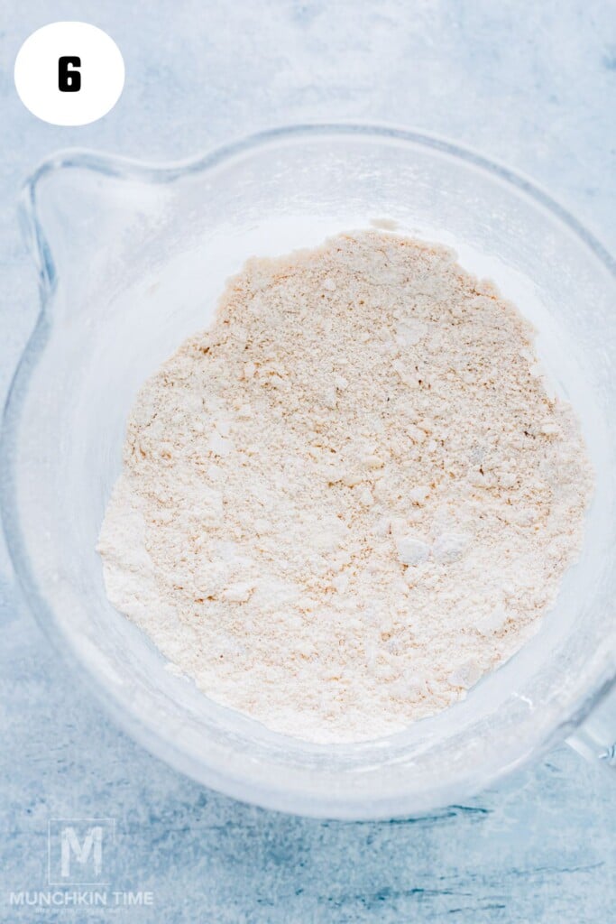 A glass mixing bowl filled with dry flour mixture sits on a light surface, with a number 6 in a white circle at the top left corner.