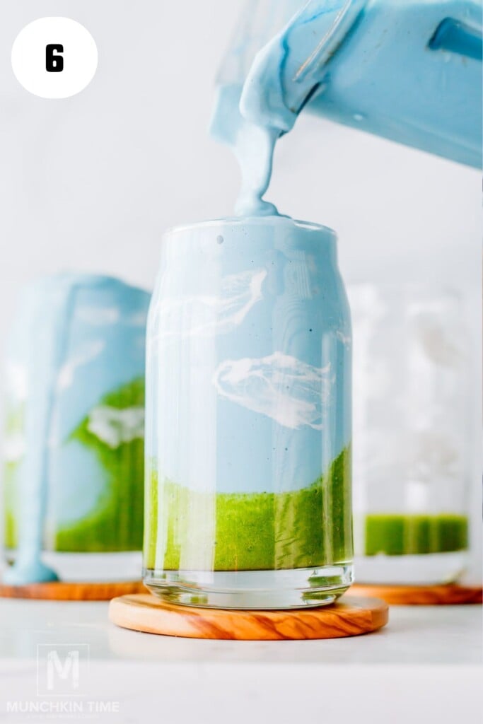A thick blue spirulina smoothie mixture is being poured from a blender into a glass with a green layer at the bottom, resembling a landscape scene.