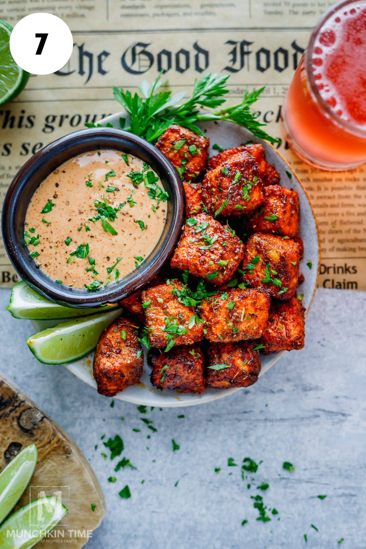 A plate of seasoned air fryer salmon bites garnished with herbs, served with lime wedges and a dipping sauce, next to a glass of pink beverage on a newspaper background.