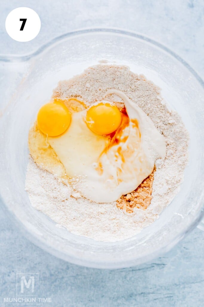 A glass bowl containing flour, two cracked eggs, vanilla extract, and wet ingredients, ready to be mixed for baking. A white circle with the number 7 is in the top left corner.