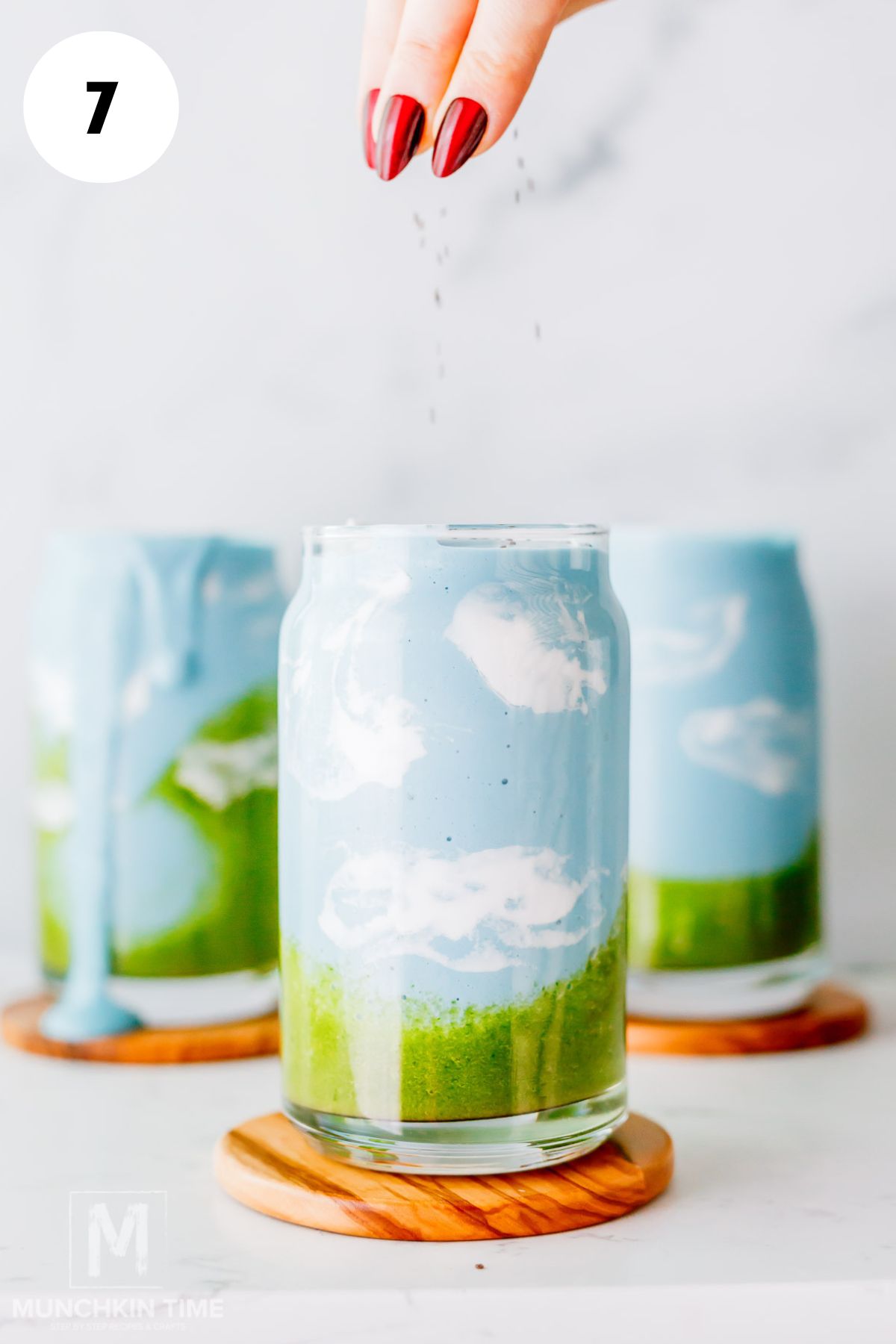 A hand sprinkles chia seeds over a layered blue and green spirulina smoothie in a glass painted to resemble a sky with clouds; two similar glasses are in the background.