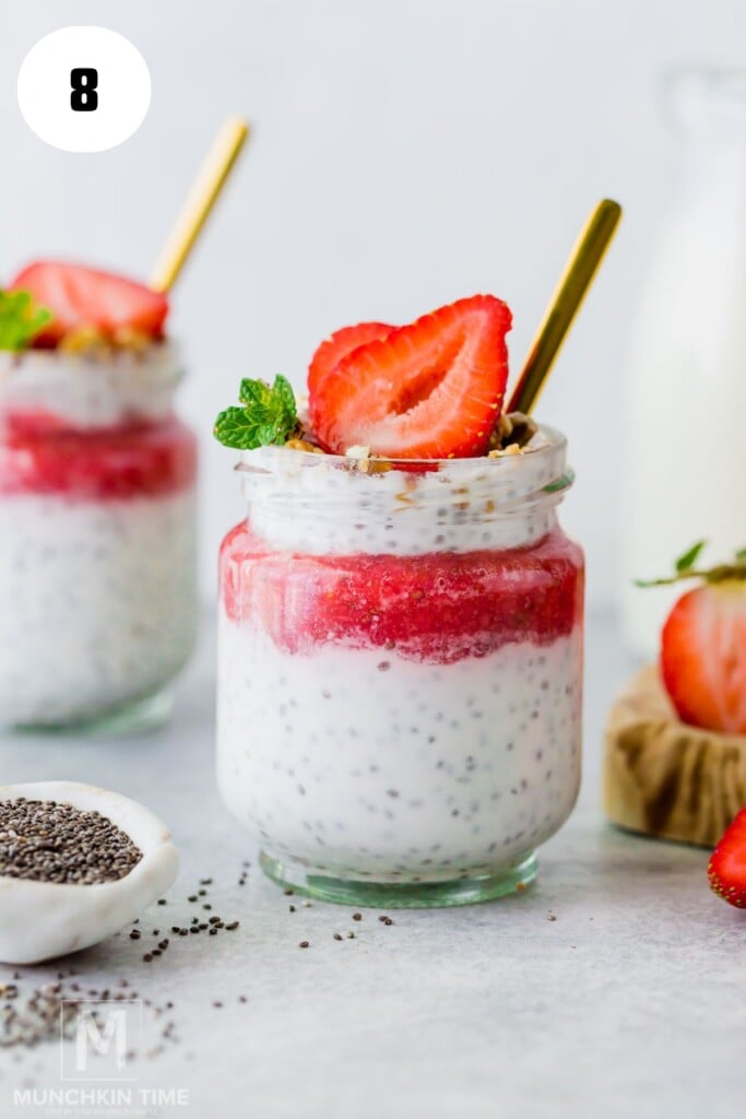 A jar of chia pudding topped with a layer of strawberry puree, fresh strawberry slices, mint leaves, and a gold spoon, with another jar and chia seeds in the background.
