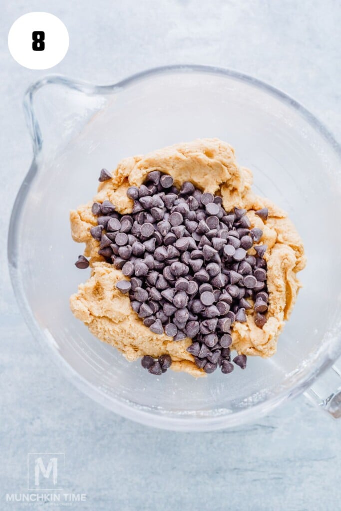 A glass mixing bowl with cookie dough and chocolate chips on top, ready to be mixed.