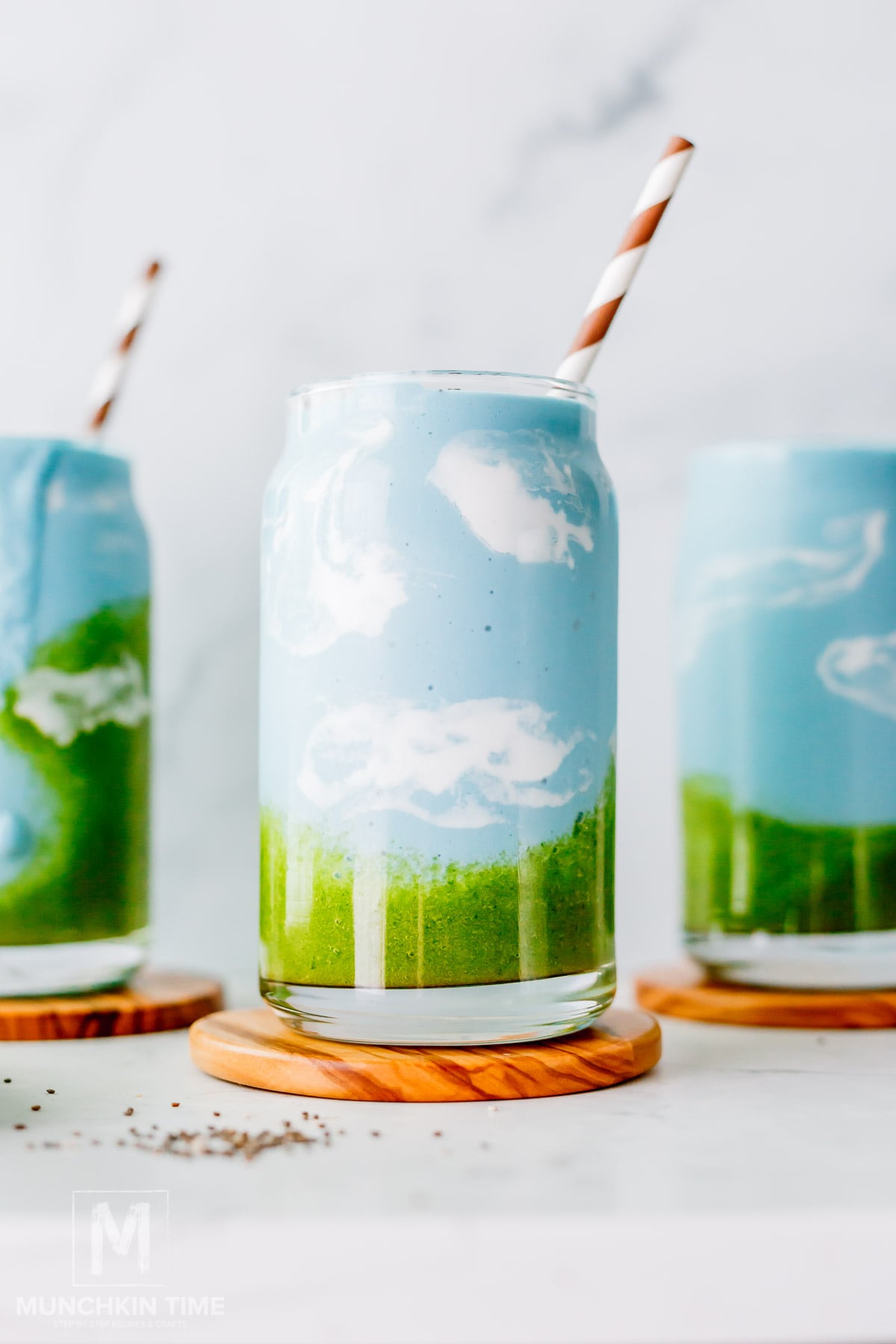Three blue spirulina smoothies in clear cans with green and blue layers, resembling a landscape with clouds, served with striped paper straws on wooden coasters.