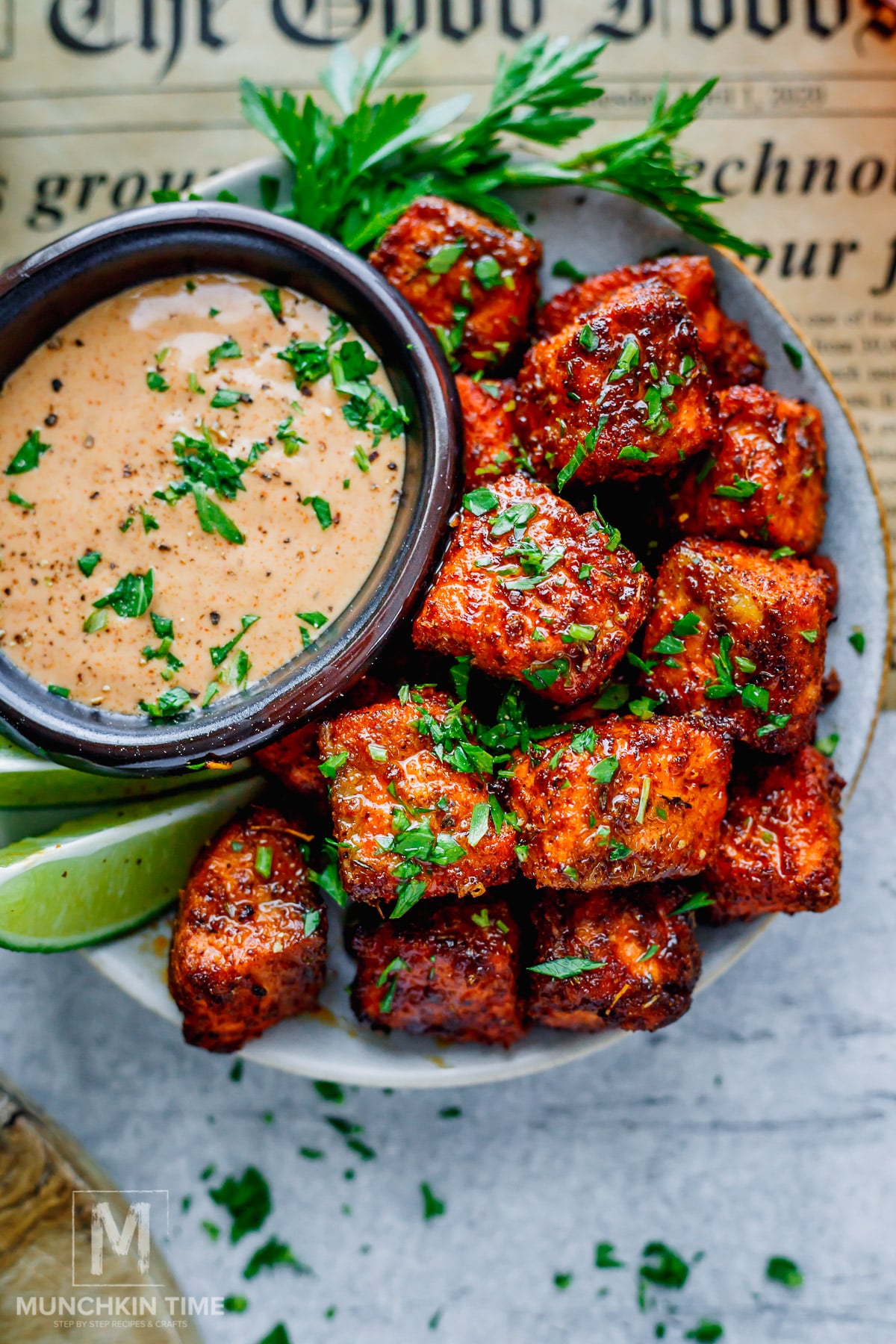 A bowl of seasoned, grilled salmon bites garnished with chopped herbs, served with a creamy dipping sauce, lime wedges, and parsley on the side.
