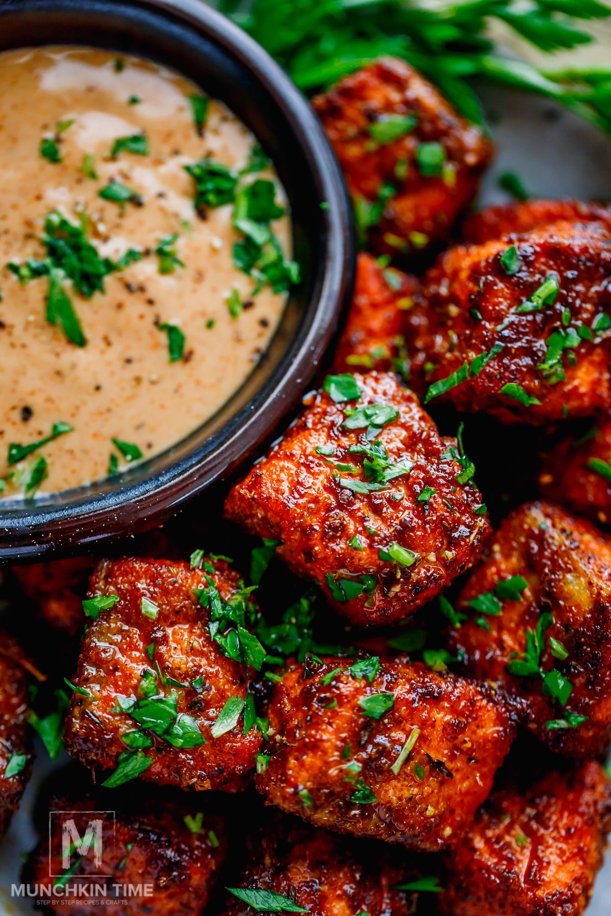 Close-up of crispy, seasoned salmon bites garnished with chopped herbs, served with a bowl of creamy dipping sauce.