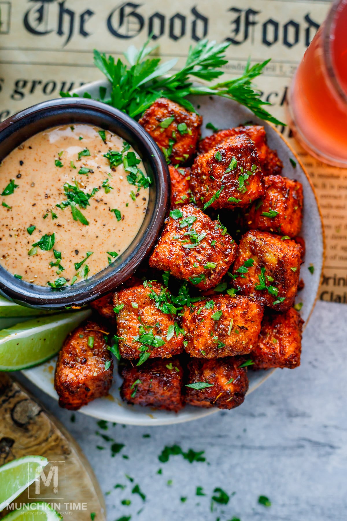 A plate of seasoned, golden-brown air fryer salmon bites garnished with herbs, served with a creamy dipping sauce, lime wedges, and a drink.