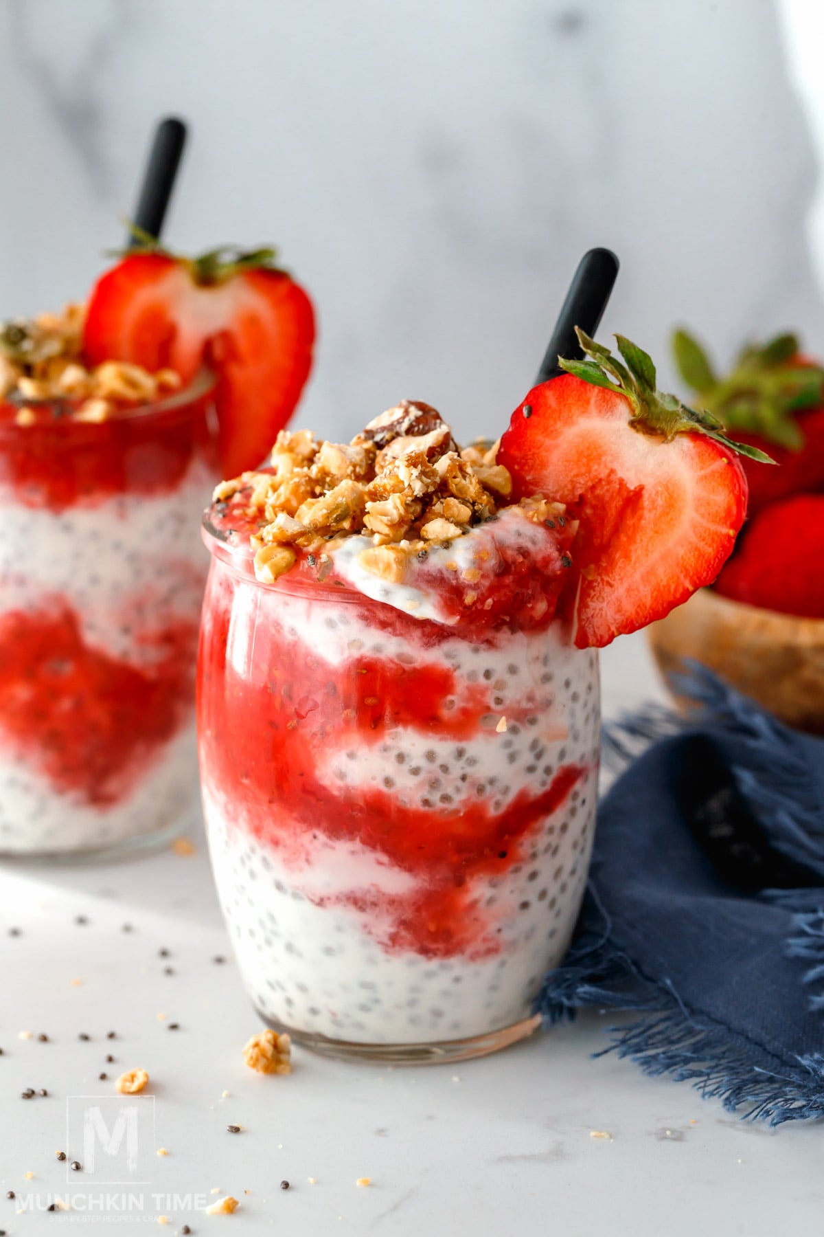 A glass of chia pudding layered with yogurt and strawberry sauce, topped with granola and a fresh strawberry slice, with a straw inserted. Another serving is in the background.