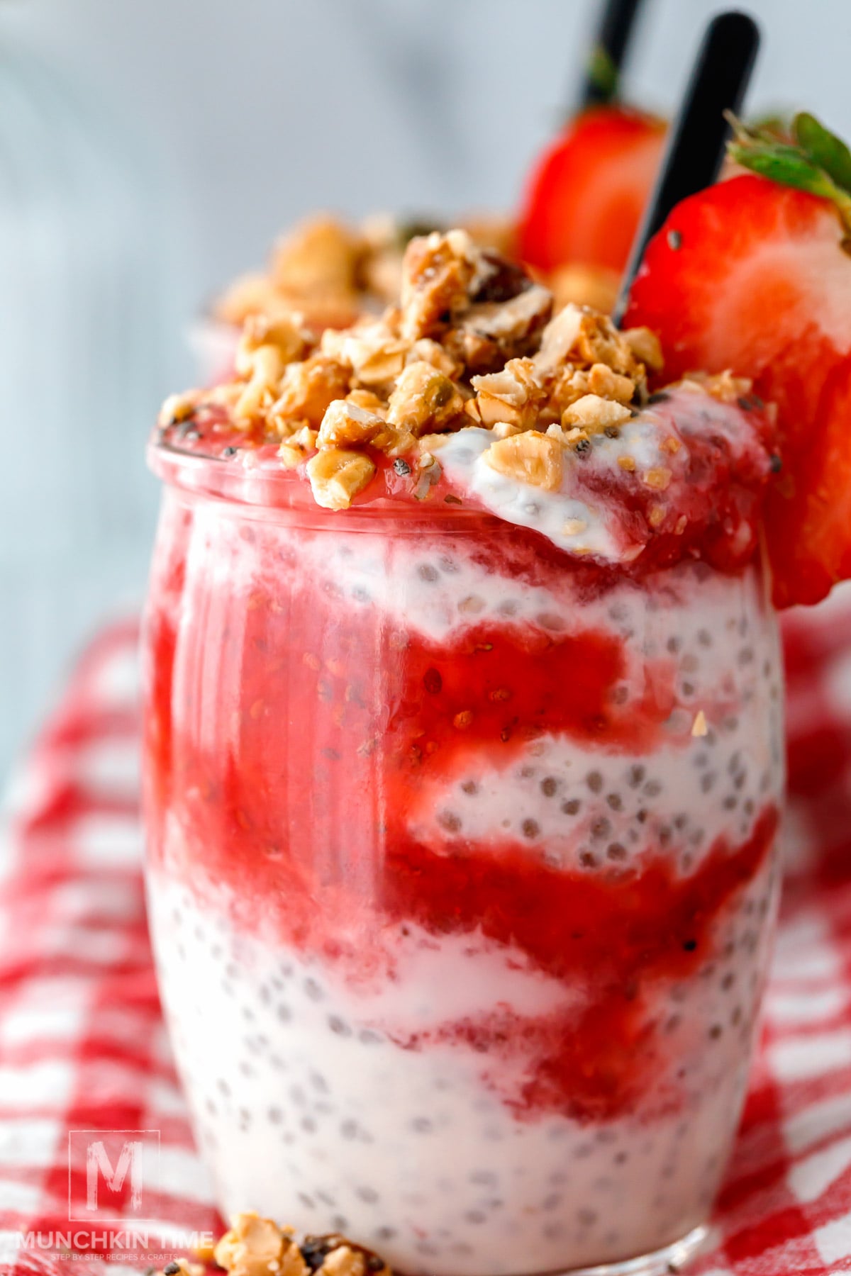 A glass filled with chia pudding layered with strawberry sauce, topped with granola and a fresh strawberry, set on a red and white checkered cloth.