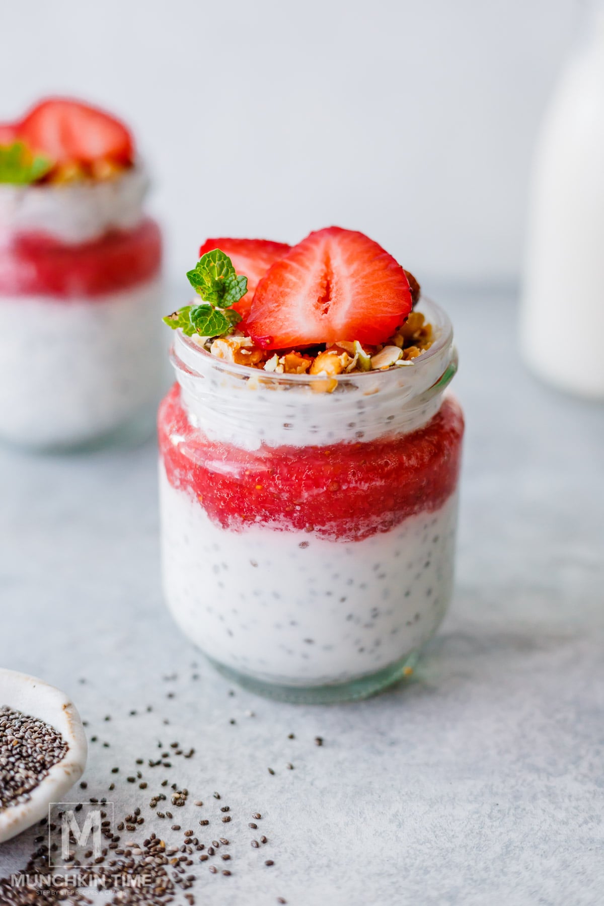A jar of chia pudding layered with strawberry puree, granola, and topped with sliced strawberries and mint on a light surface.