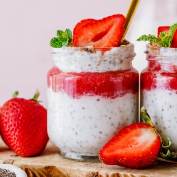 A jar of chia pudding topped with strawberry puree, a sliced strawberry, and mint leaves, placed on a wooden board with fresh strawberries nearby.