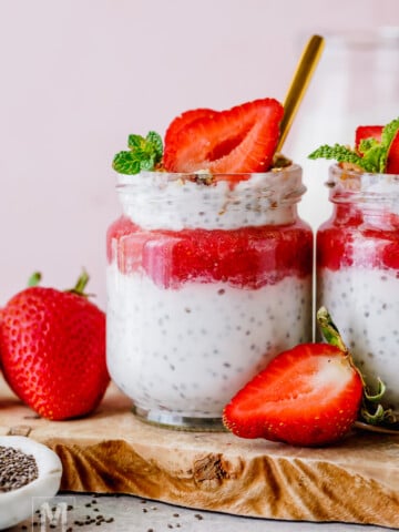 A jar of chia pudding topped with strawberry puree, a sliced strawberry, and mint leaves, placed on a wooden board with fresh strawberries nearby.