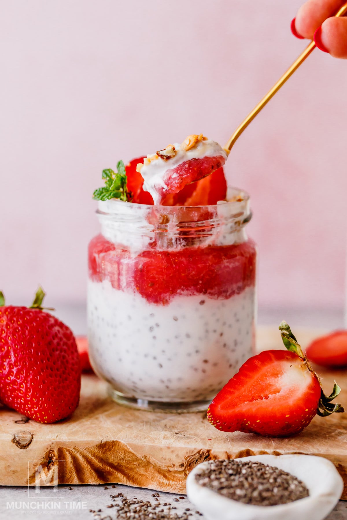 A glass jar filled with chia pudding, yogurt, and sliced strawberries, topped with granola and mint, with a spoon lifting out a portion. Fresh strawberries and chia seeds are beside the jar.