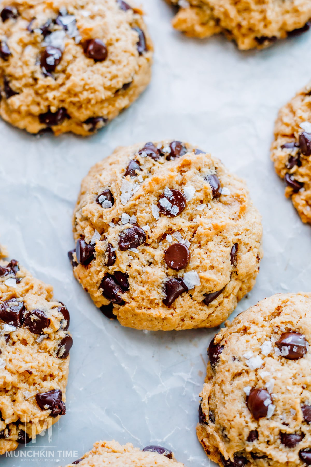 Close-up of several sourdough chocolate chip cookies topped with flaky sea salt, arranged on a white parchment paper background.
