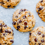 Close-up of several sourdough chocolate chip cookies sprinkled with coarse sea salt, arranged on a light-colored surface.