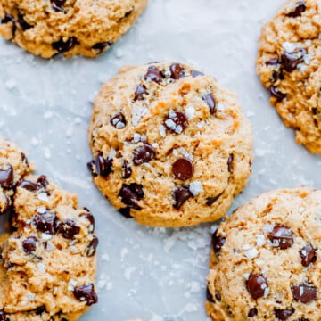 Close-up of several sourdough chocolate chip cookies sprinkled with coarse sea salt, arranged on a light-colored surface.