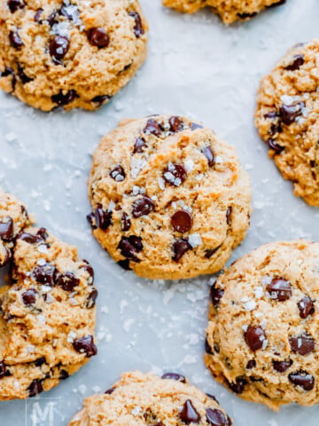 Close-up of several sourdough chocolate chip cookies sprinkled with coarse sea salt, arranged on a light-colored surface.
