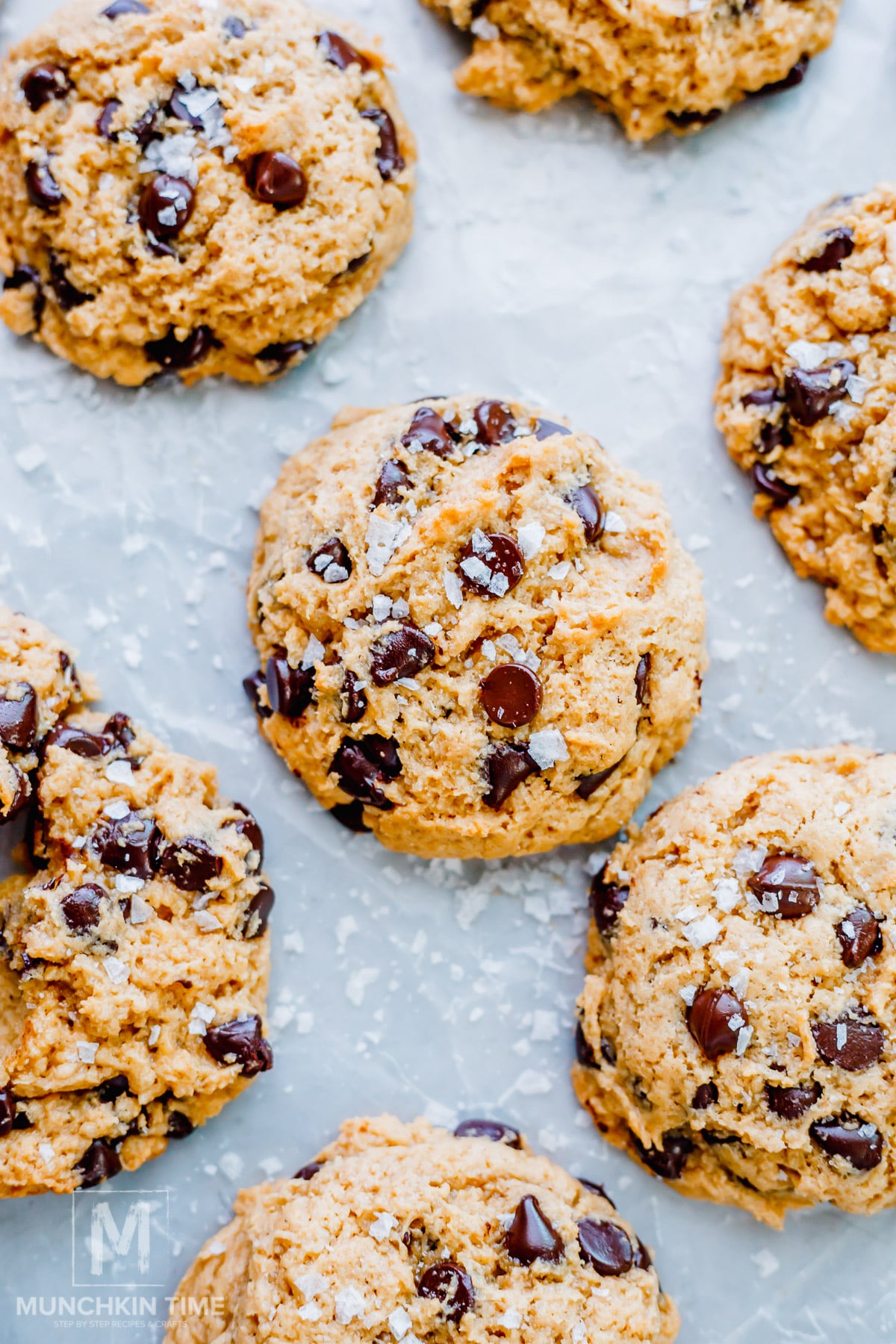 Close-up of several sourdough chocolate chip cookies sprinkled with coarse sea salt, arranged on a light-colored surface.