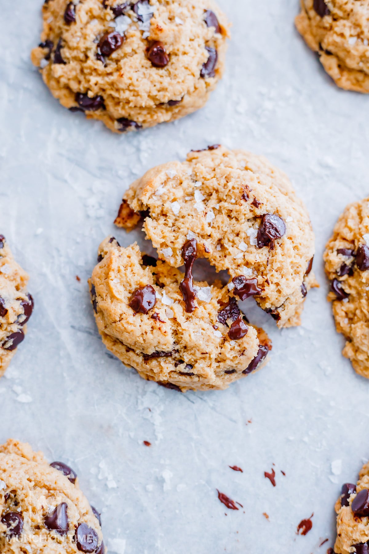 A close-up of sourdough chocolate chip cookies on parchment paper, with one cookie broken in half showing the melted chocolate inside.