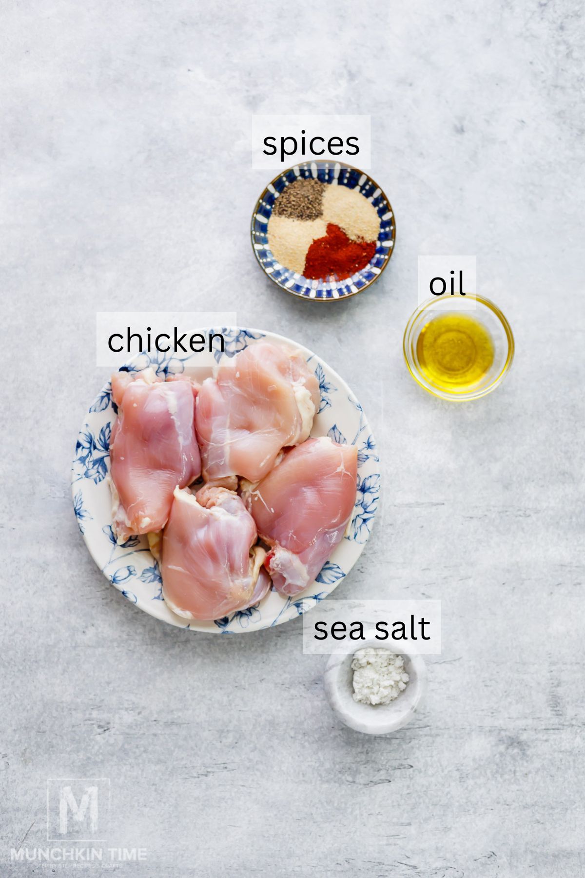 Raw chicken thighs on a floral plate, a small bowl of mixed spices, a glass cup of oil, and a dish of sea salt arranged on a light surface, each labeled.
