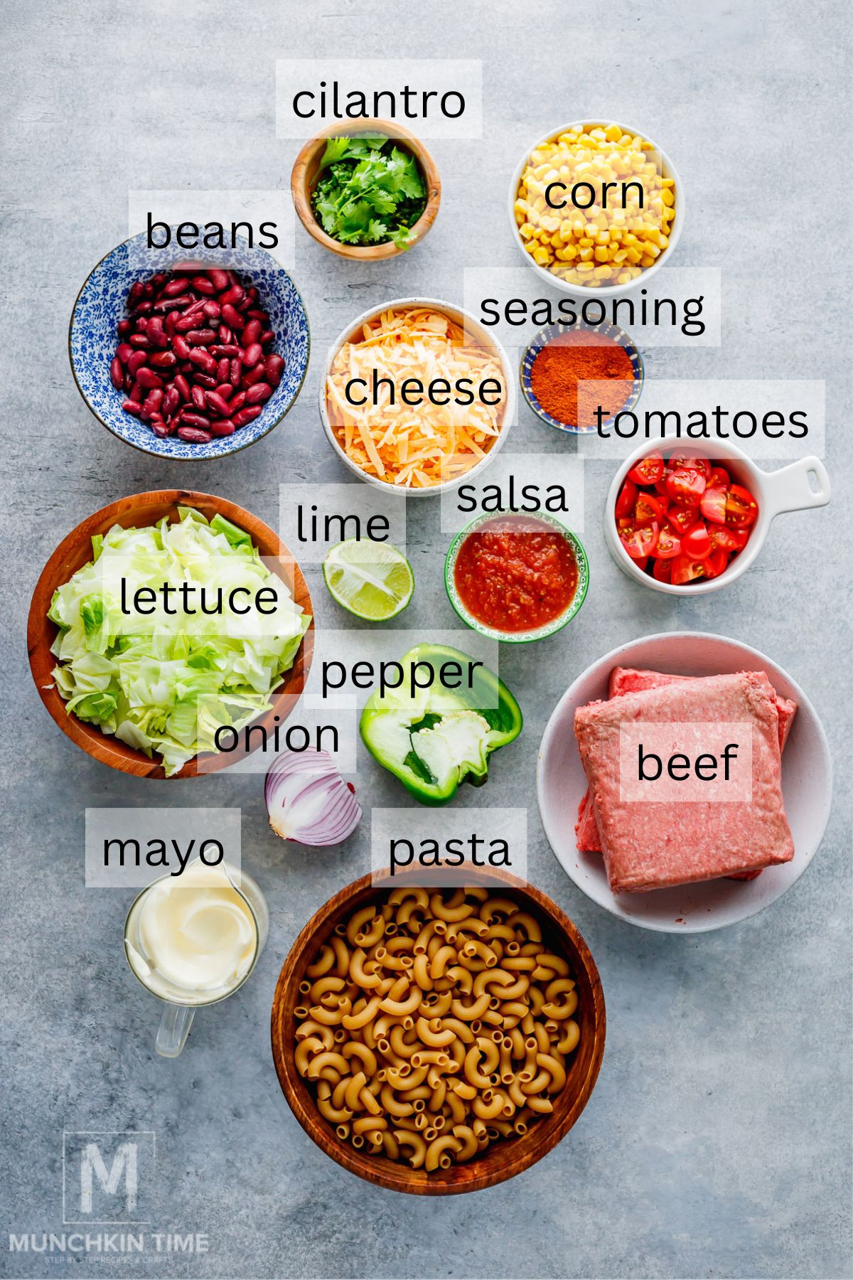 A variety of labeled ingredients including ground beef, pasta, beans, cheese, lettuce, corn, tomatoes, salsa, lime, onion, pepper, cilantro, mayo, and seasoning on a countertop.