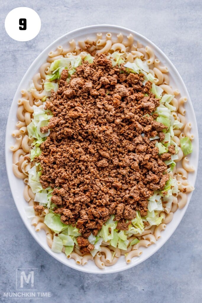 An oval dish filled with cooked elbow macaroni, chopped lettuce, and seasoned ground beef, layered in that order. A number 9 is in a white circle in the top left corner.