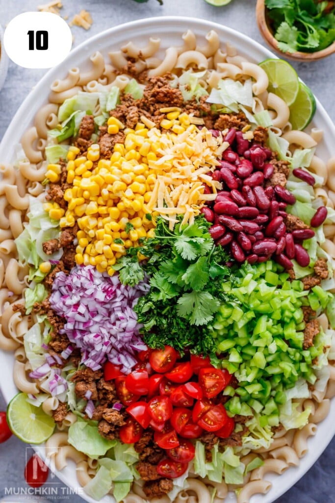 An overhead view of a salad with macaroni, ground meat, kidney beans, corn, shredded cheese, chopped lettuce, tomatoes, red onion, green bell pepper, and cilantro on a white platter.