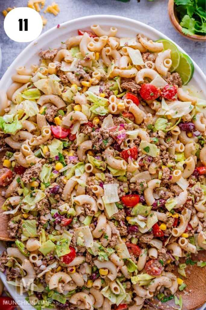 A large plate of taco pasta salad with macaroni, ground beef, lettuce, tomatoes, corn, and beans, garnished with chopped herbs.