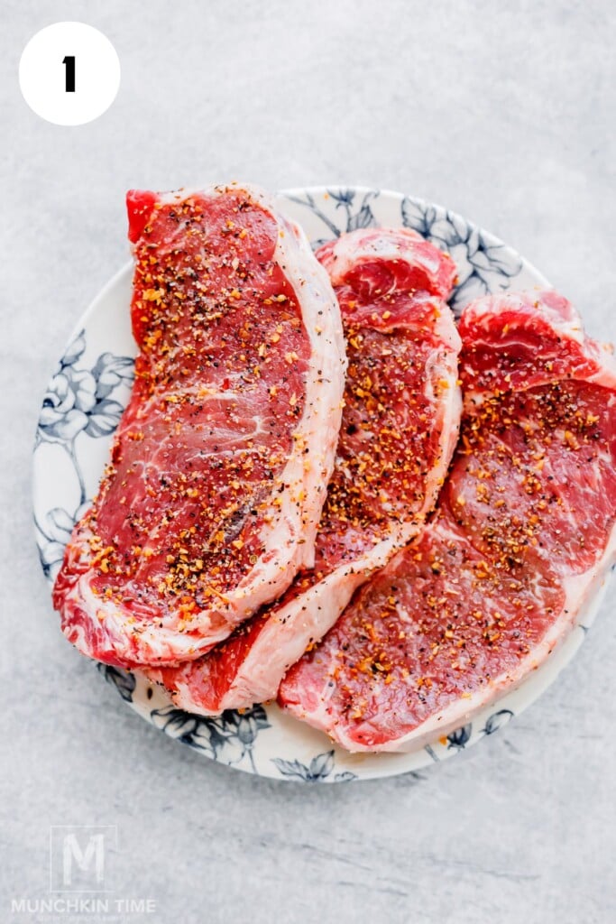 Three raw beef steaks seasoned with pepper and spices on a white plate with a floral design, placed on a light-colored surface.