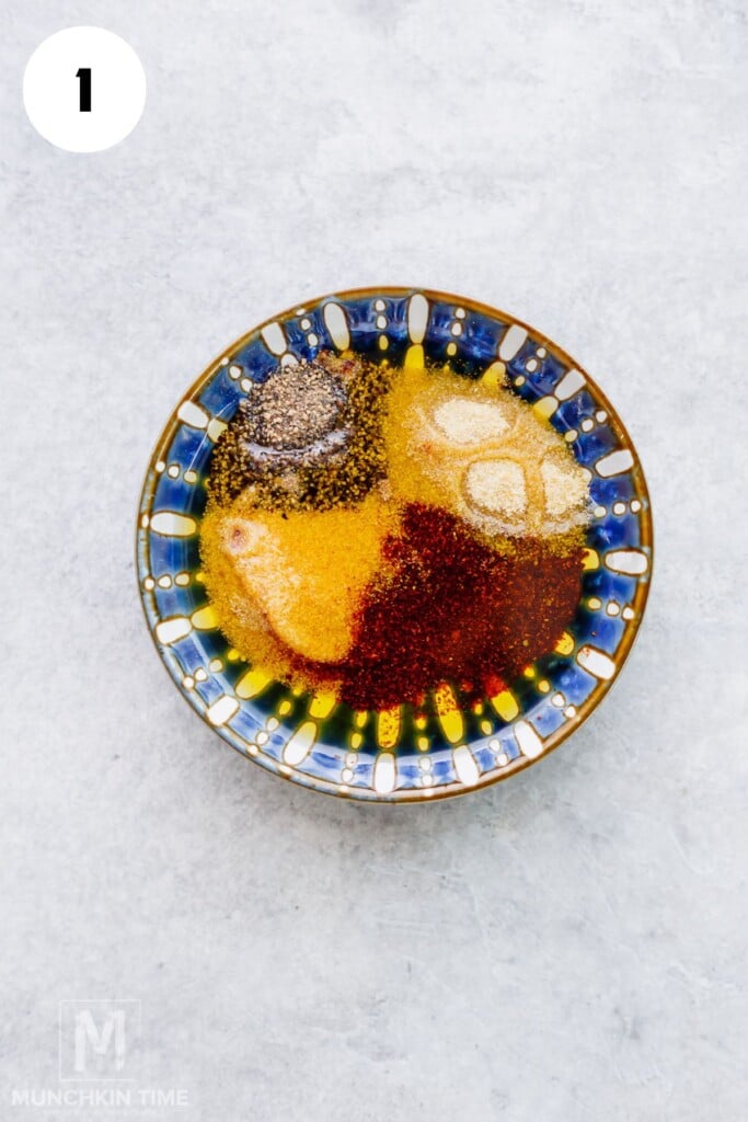 A patterned bowl containing oil and various ground spices, including black pepper, garlic powder, and paprika, on a light gray surface.