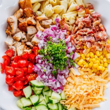 On a large serving plate chopping cooked chicken, bacon, cucumber, tomatoes, onion, and cheddar cheese for pasta salad.