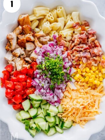 On a large serving plate chopping cooked chicken, bacon, cucumber, tomatoes, onion, and cheddar cheese for pasta salad.