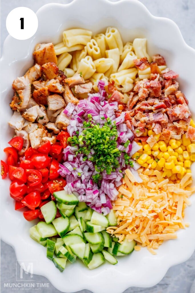 On a large serving plate chopping cooked chicken, bacon, cucumber, tomatoes, onion, and cheddar cheese for pasta salad.