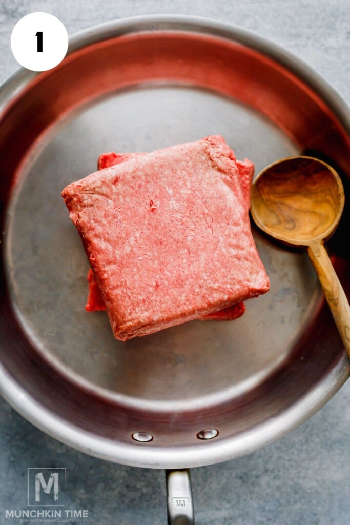 Two blocks of raw ground beef in a stainless steel pan with a wooden spoon on the side.