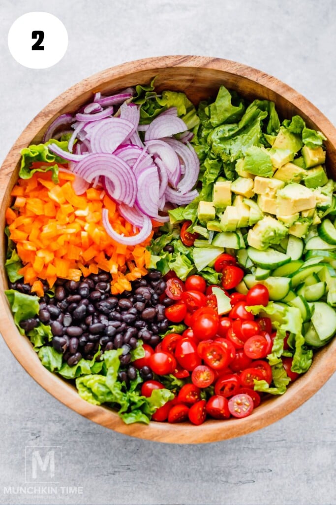 A wooden bowl filled with chopped lettuce, sliced red onion, diced bell pepper, black beans, cherry tomatoes, cucumber, and avocado, arranged in sections.