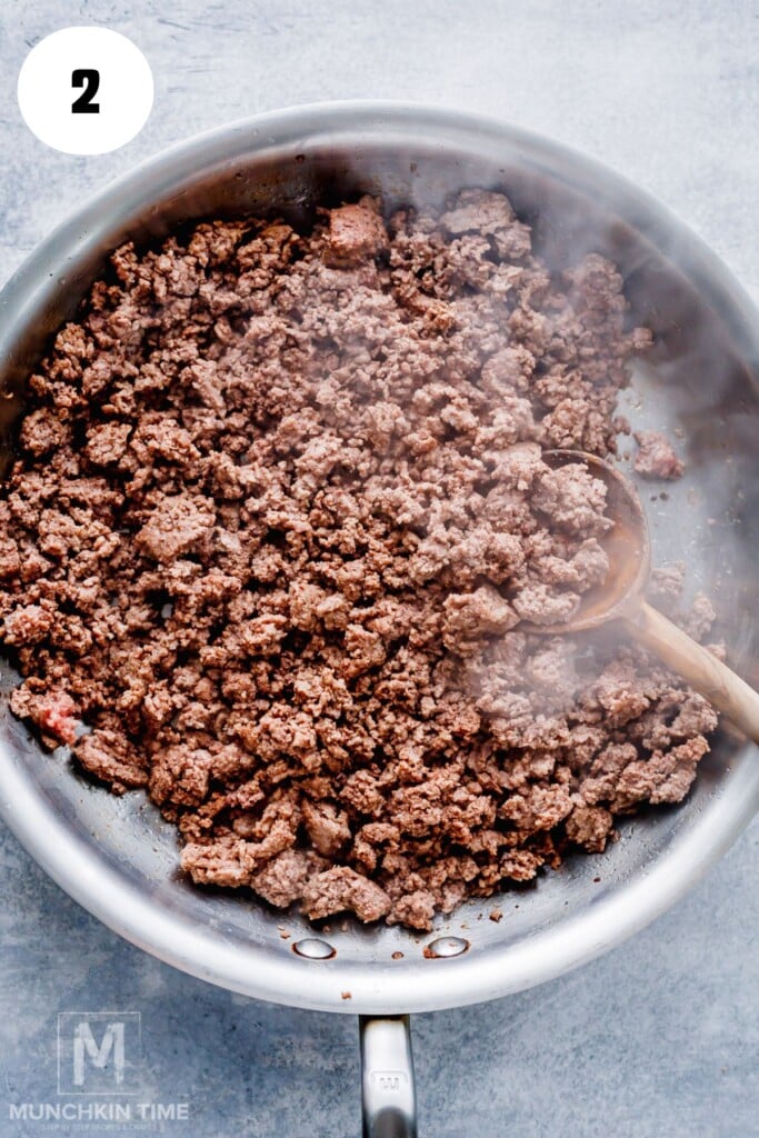 Ground beef being browned in a stainless steel skillet with a wooden spoon, viewed from above. Steam is visible rising from the meat.