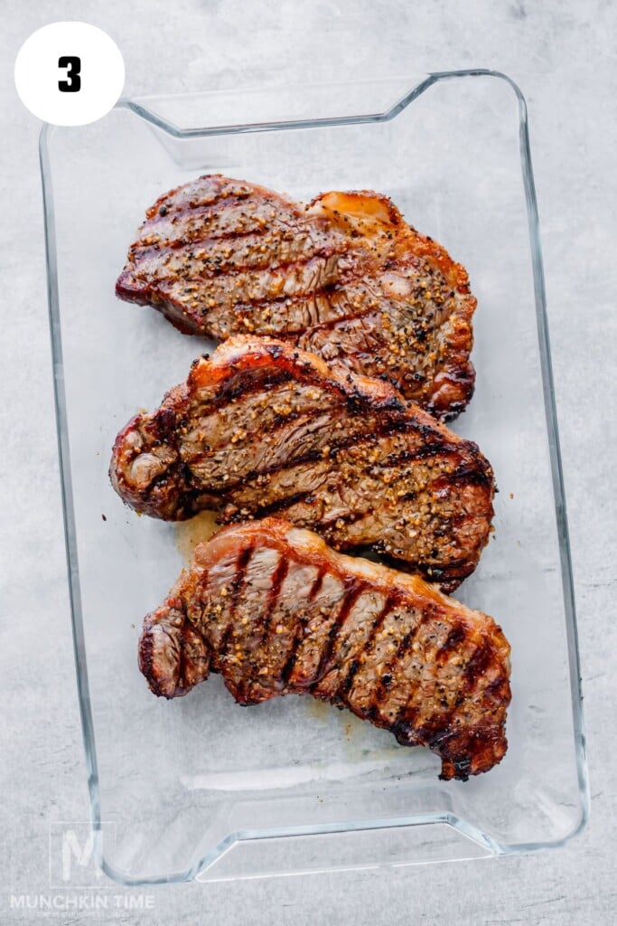 Three cooked, seasoned steaks are arranged in a clear rectangular glass dish on a light gray surface.