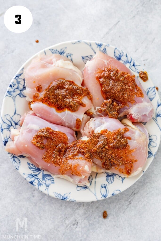 Four raw chicken thighs on a white and blue floral plate, topped with a reddish-brown spice mixture, placed on a light gray surface.