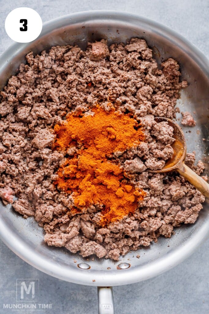 Ground beef cooking in a pan with taco seasoning sprinkled on top and a wooden spoon on the side.