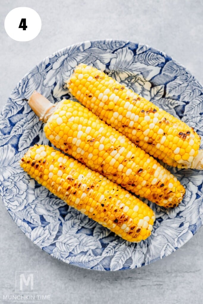 Three grilled corn on the cob are placed in a blue and white patterned bowl on a light surface.