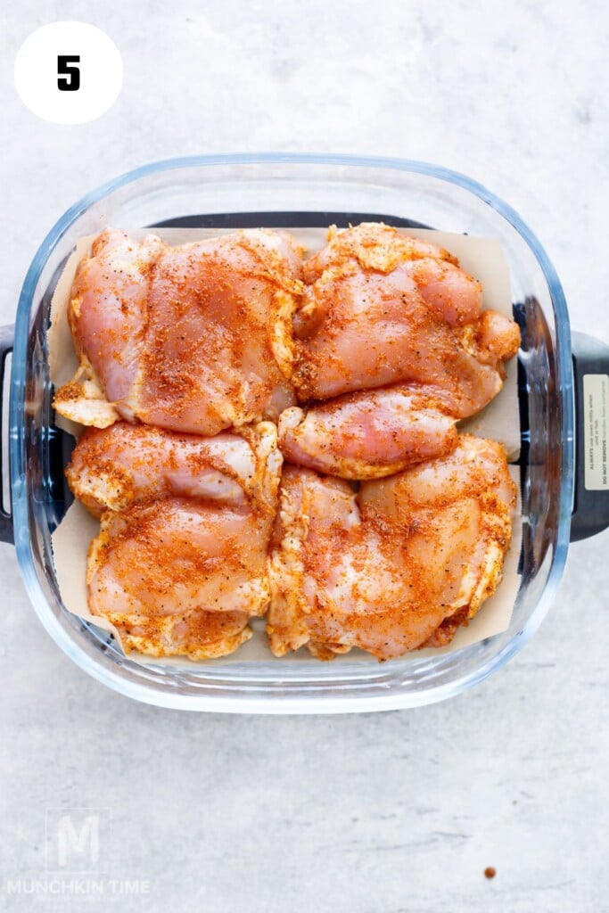 Four seasoned raw chicken thighs are arranged in a square glass baking dish on a light-colored surface.