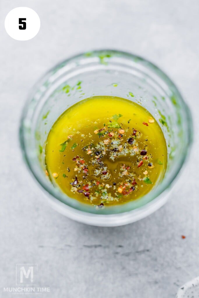 A glass jar filled with olive oil, herbs, and spices, viewed from above. The number 5 is in a white circle in the top left corner.