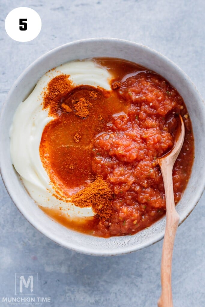 A bowl with sour cream, red tomato salsa, and ground spices being mixed together with a wooden spoon.