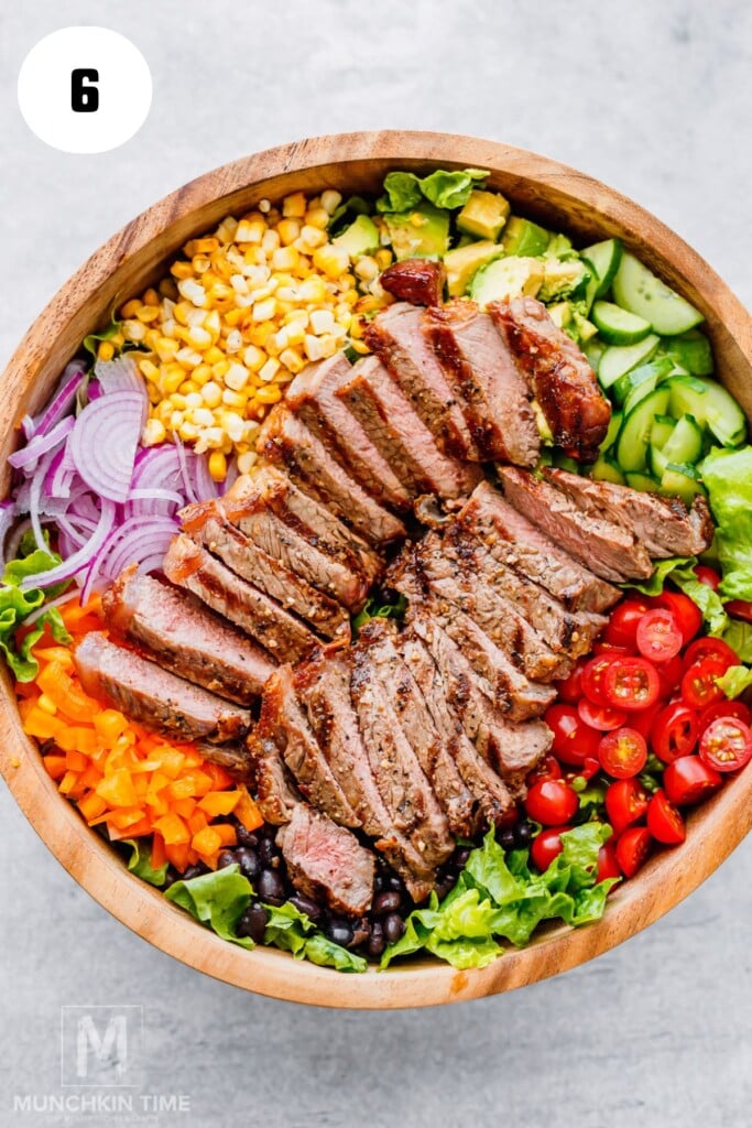 A wooden bowl filled with sliced grilled steak, cherry tomatoes, black beans, red onion, corn, bell pepper, cucumber, and lettuce arranged in sections.