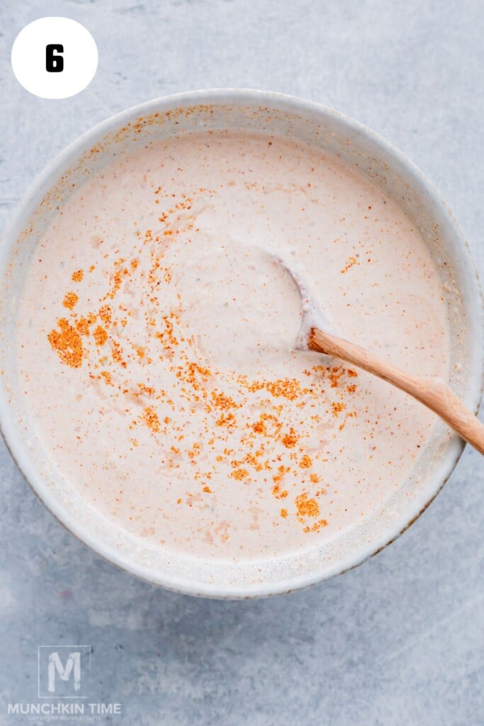 Bowl of creamy sauce with orange spices sprinkled on top, being stirred with a wooden spoon. A number 6 is in the top left corner.