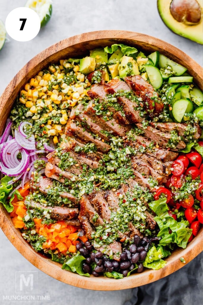 Wooden bowl filled with sliced grilled steak, black beans, corn, cherry tomatoes, cucumbers, red onion, bell pepper, avocado, lettuce, and chimichurri sauce.