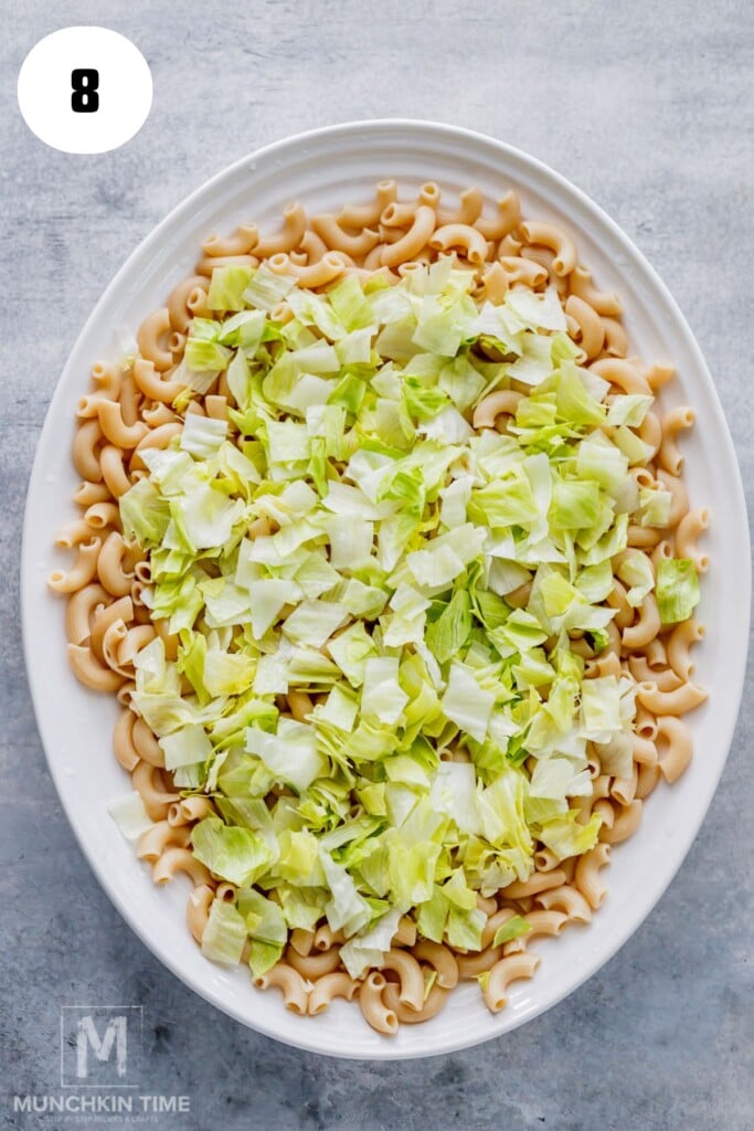 Oval white dish filled with cooked elbow macaroni topped with chopped iceberg lettuce on a gray surface. Number 8 in a white circle appears in the top left corner.
