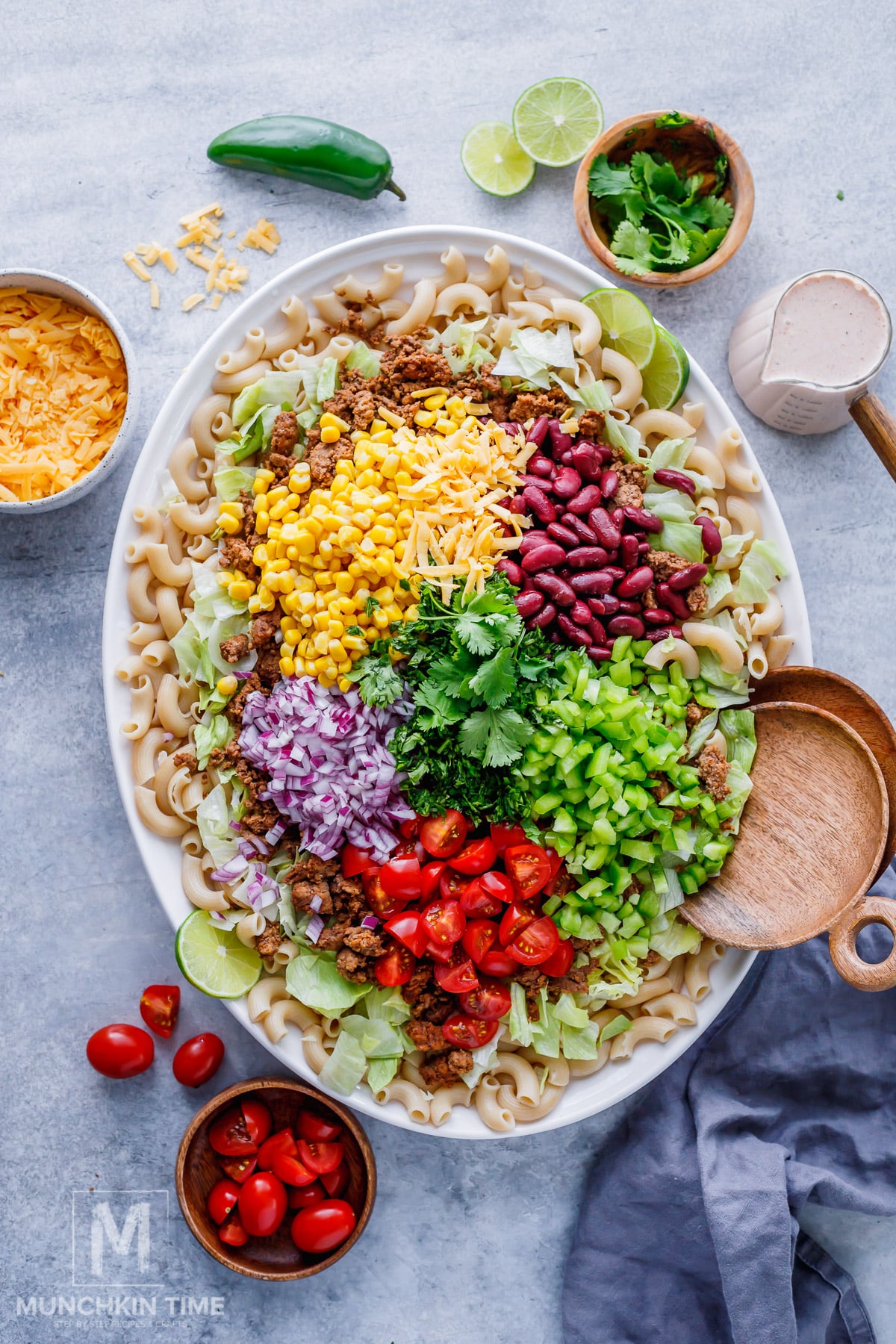 A large oval platter with macaroni pasta topped with ground meat, shredded cheese, corn, kidney beans, chopped lettuce, tomatoes, red onion, celery, and cilantro. Surrounding bowls hold toppings.