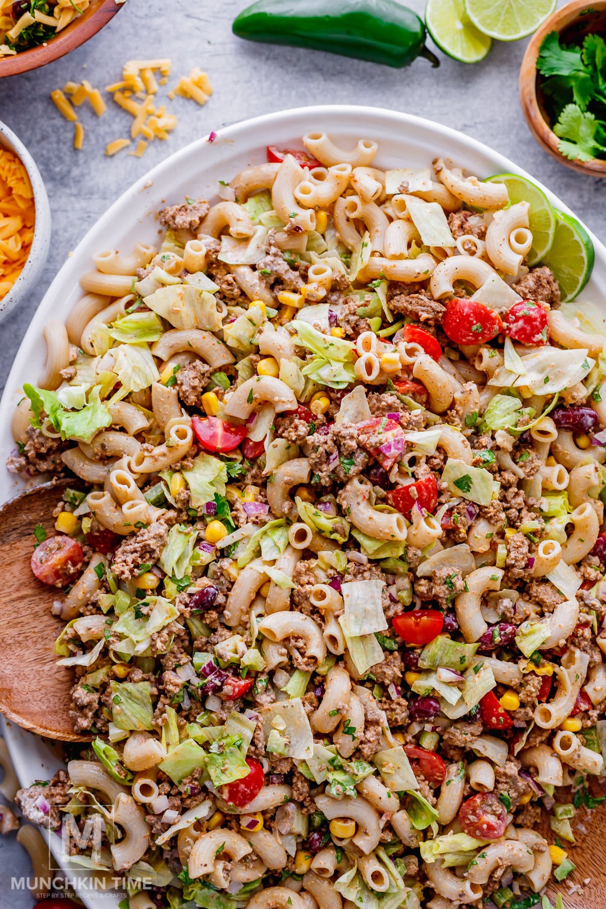 A large plate of taco pasta salad with ground beef, lettuce, tomatoes, corn, and macaroni, surrounded by bowls of shredded cheese, cilantro, and lime wedges.